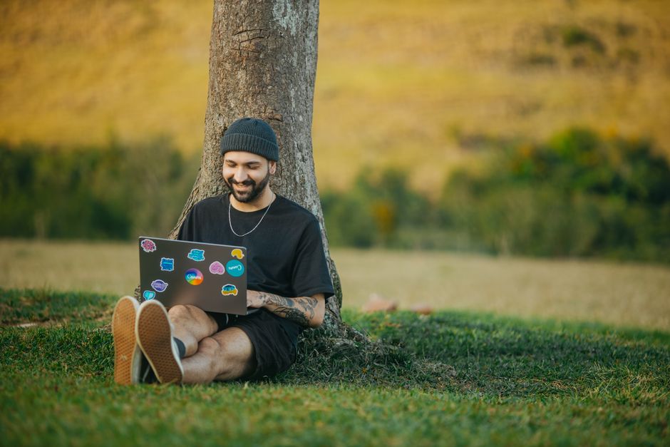 Bărbat la laptop într-o cameră luminoasă, cu un pahar de apă și un ventilator pe birou